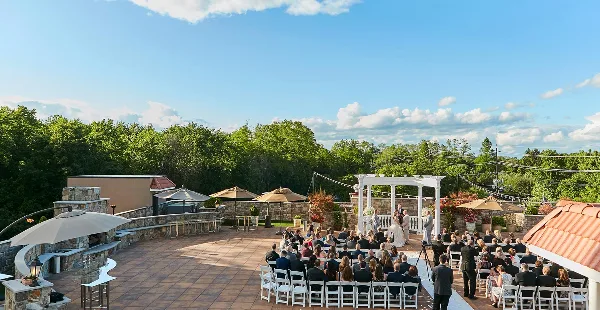 A wedding ceremony in progress on a rooftop terrace with a white arch, seated guests, and a view of trees and sky.