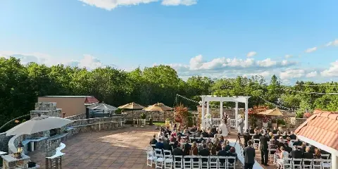 A wedding ceremony in progress on a rooftop terrace with a white arch, seated guests, and a view of trees and sky.