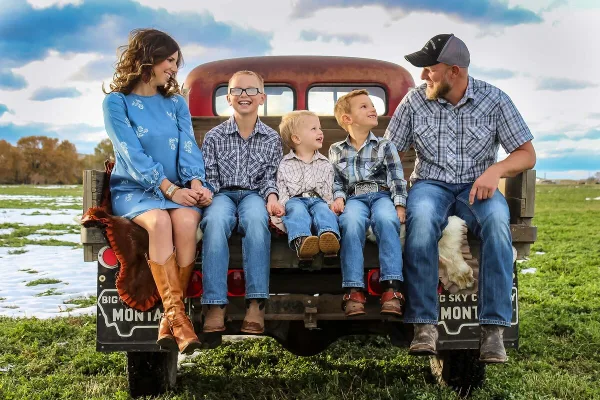 A family of five sitting on the tailgate of a vintage truck in a Montana field with snow patches and a blue sky.