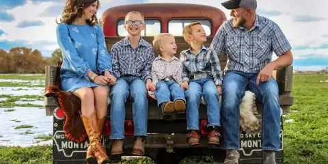 A family of five sitting on the tailgate of a vintage truck in a Montana field with snow patches and a blue sky.