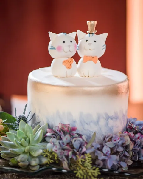 A white and gold ombre cake topped with two sculpted white cat figures, one wearing a top hat and the other holding a small bouquet, surrounded by succulents and hydrangeas.