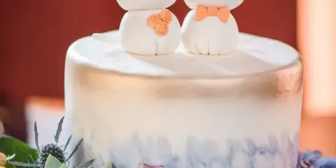 A white and gold ombre cake topped with two sculpted white cat figures, one wearing a top hat and the other holding a small bouquet, surrounded by succulents and hydrangeas.