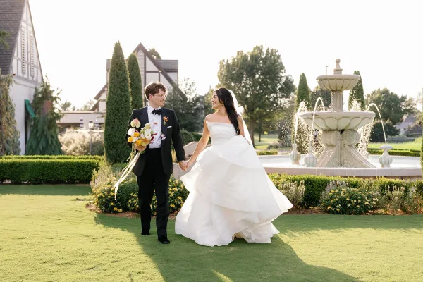 A bride and groom holding hands and walking across a manicured lawn in front of a stone fountain and a Tudor-style building.