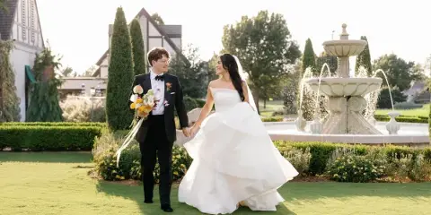 A bride and groom holding hands and walking across a manicured lawn in front of a stone fountain and a Tudor-style building.