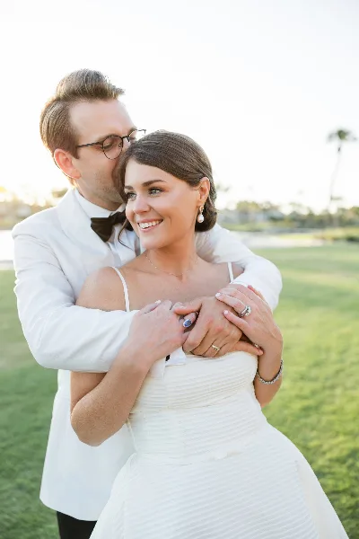 A bride and groom in white formal attire embracing outdoors on a grassy field.