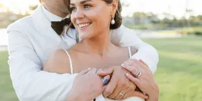 A bride and groom in white formal attire embracing outdoors on a grassy field.