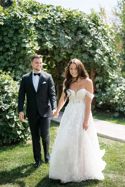 A bride and groom holding hands and walking on a grassy lawn with a leafy green hedge in the background.