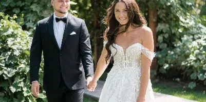A bride and groom holding hands and walking on a grassy lawn with a leafy green hedge in the background.