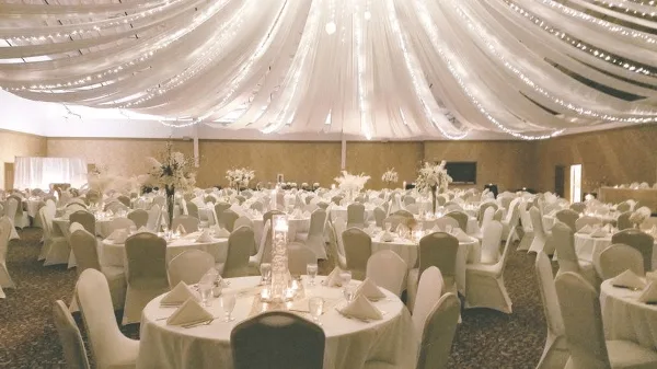 A spacious event venue featuring round tables with white linens, floral centerpieces, and a ceiling decorated with draped fabric and string lights.