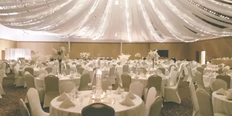 A spacious event venue featuring round tables with white linens, floral centerpieces, and a ceiling decorated with draped fabric and string lights.