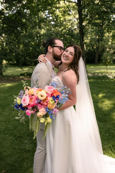 A bride and groom embracing outdoors, with the bride holding a colorful bouquet of flowers.