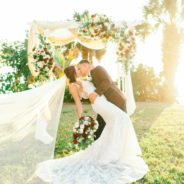 A bride and groom kissing under a floral wedding arch with a bouquet of red and white roses.