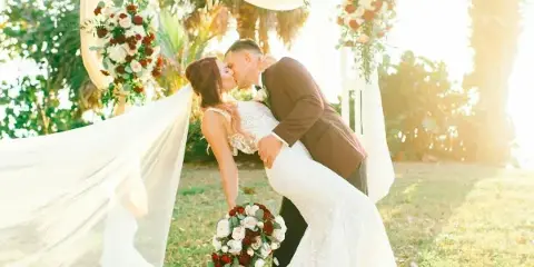 A bride and groom kissing under a floral wedding arch with a bouquet of red and white roses.