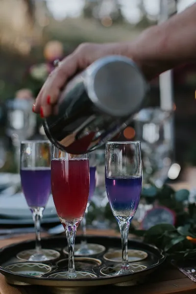 A bartender pouring a colorful cocktail from a shaker into a champagne flute at an outdoor event.
