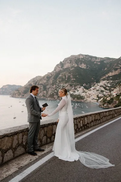 A bride and groom exchanging vows on a coastal road with a scenic mountain and sea background.
