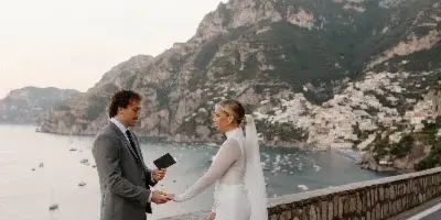 A bride and groom exchanging vows on a coastal road with a scenic mountain and sea background.