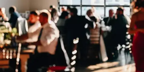 A server in a white shirt and suspenders attends to guests at a formal event with tables and floral arrangements.
