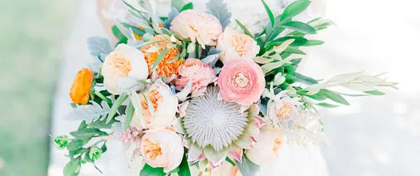 A close-up photograph of a wedding bouquet featuring peach ranunculus, a white protea, and greenery.