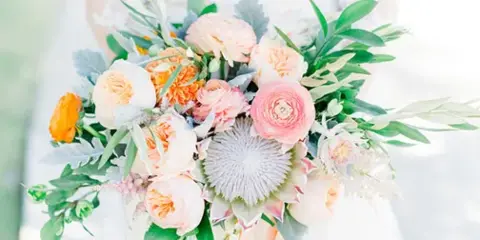 A close-up photograph of a wedding bouquet featuring peach ranunculus, a white protea, and greenery.