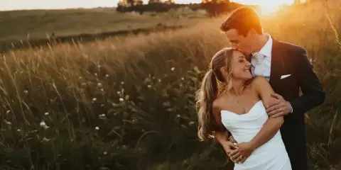 A bride and groom embracing in a field of tall grass during golden hour.