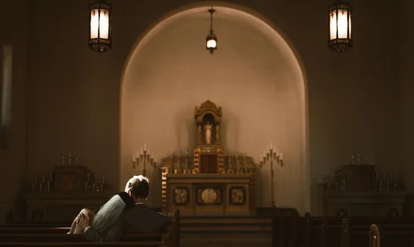 A cinematic photograph of a couple embracing in a church pew, facing an ornate altar with warm lighting.