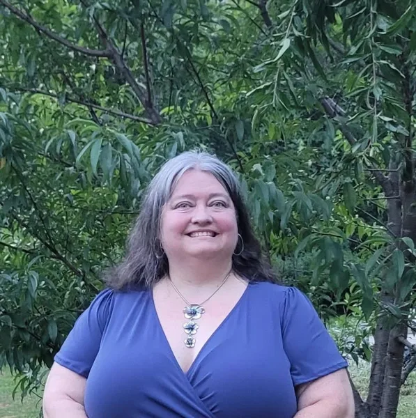 Portrait of a smiling woman with gray-streaked hair wearing a blue top and necklace, standing outdoors in front of green foliage.