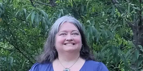 Portrait of a smiling woman with gray-streaked hair wearing a blue top and necklace, standing outdoors in front of green foliage.