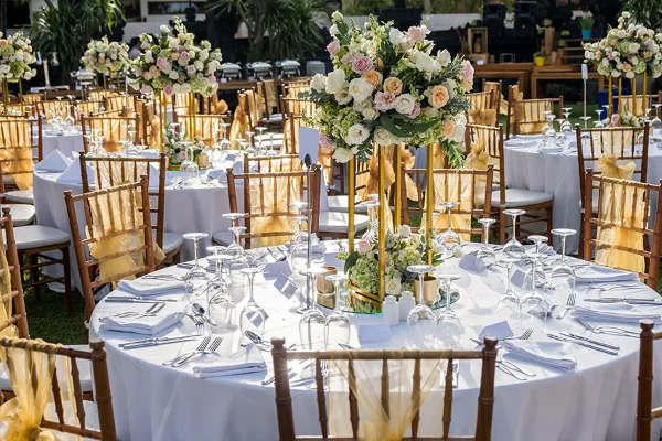 An outdoor event setup featuring round tables with white tablecloths, Chiavari chairs, and floral centerpieces.