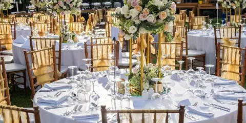 An outdoor event setup featuring round tables with white tablecloths, Chiavari chairs, and floral centerpieces.
