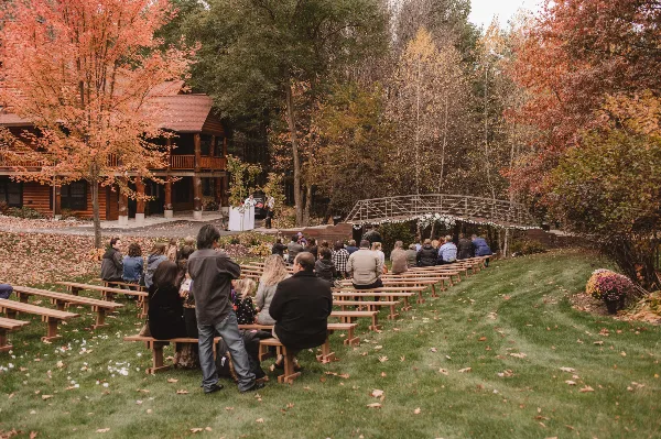 Outdoor wedding ceremony at a rustic retreat with guests seated on wooden benches facing a decorated arch and log lodge.