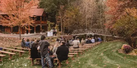 Outdoor wedding ceremony at a rustic retreat with guests seated on wooden benches facing a decorated arch and log lodge.