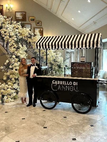 A couple in wedding attire standing next to a black and white striped cannoli cart at a wedding venue.