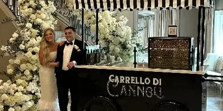 A couple in wedding attire standing next to a black and white striped cannoli cart at a wedding venue.