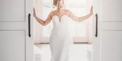 A bride in a white wedding dress standing in a doorway with her arms outstretched.