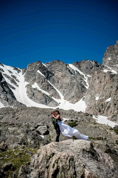 A bride and groom embracing on a rocky mountain summit with snow-covered peaks in the background.