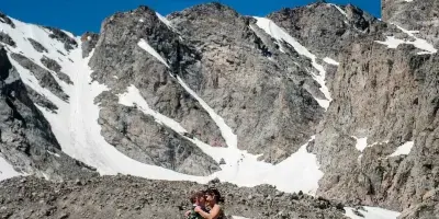 A bride and groom embracing on a rocky mountain summit with snow-covered peaks in the background.