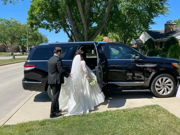 A uniformed chauffeur helps a bride in a white wedding dress step out of a black luxury SUV on a residential street.