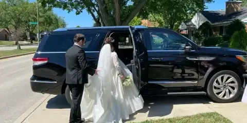 A uniformed chauffeur helps a bride in a white wedding dress step out of a black luxury SUV on a residential street.