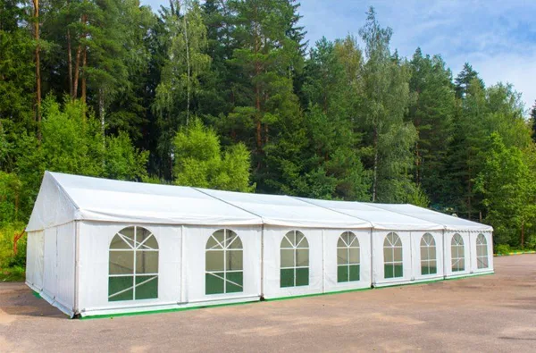 A large white event tent with arched windows set up outdoors in front of a forest.