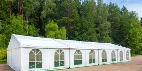 A large white event tent with arched windows set up outdoors in front of a forest.