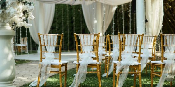 A wedding ceremony setup featuring bamboo chiavari chairs with white sashes, a draped arch, and floral arrangements.