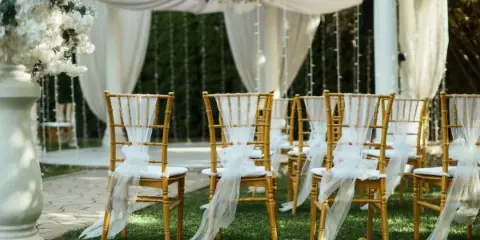 A wedding ceremony setup featuring bamboo chiavari chairs with white sashes, a draped arch, and floral arrangements.