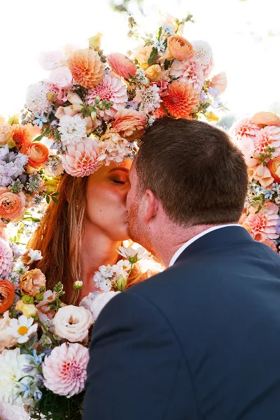 A bride and groom kissing at a wedding ceremony under a large floral arch.