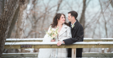 A bride and groom in a winter setting, standing near a wooden fence in a wooded area.