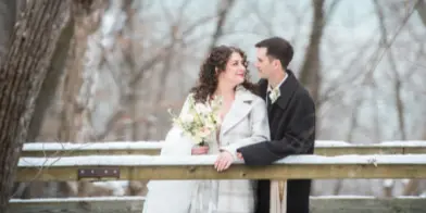 A bride and groom in a winter setting, standing near a wooden fence in a wooded area.