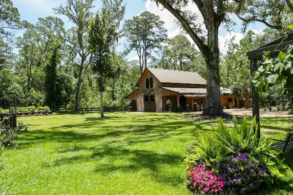 A rustic wooden barn venue surrounded by green lawns, oak trees, and flowering plants.