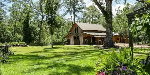 A rustic wooden barn venue surrounded by green lawns, oak trees, and flowering plants.