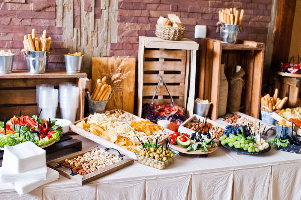 A catering buffet table featuring platters of appetizers, breadsticks, and cheeses set against a rustic brick and wood background.