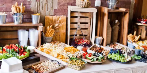 A catering buffet table featuring platters of appetizers, breadsticks, and cheeses set against a rustic brick and wood background.