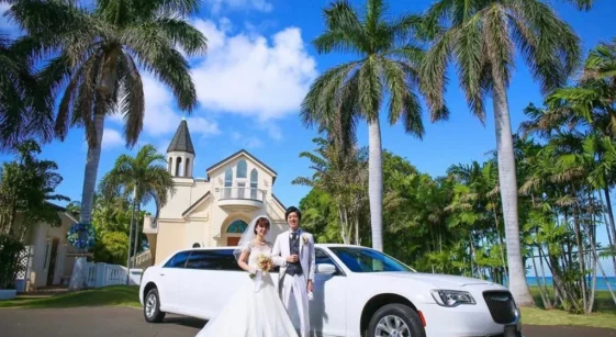 A bride and groom posing with a white luxury SUV in front of a tropical venue with palm trees.